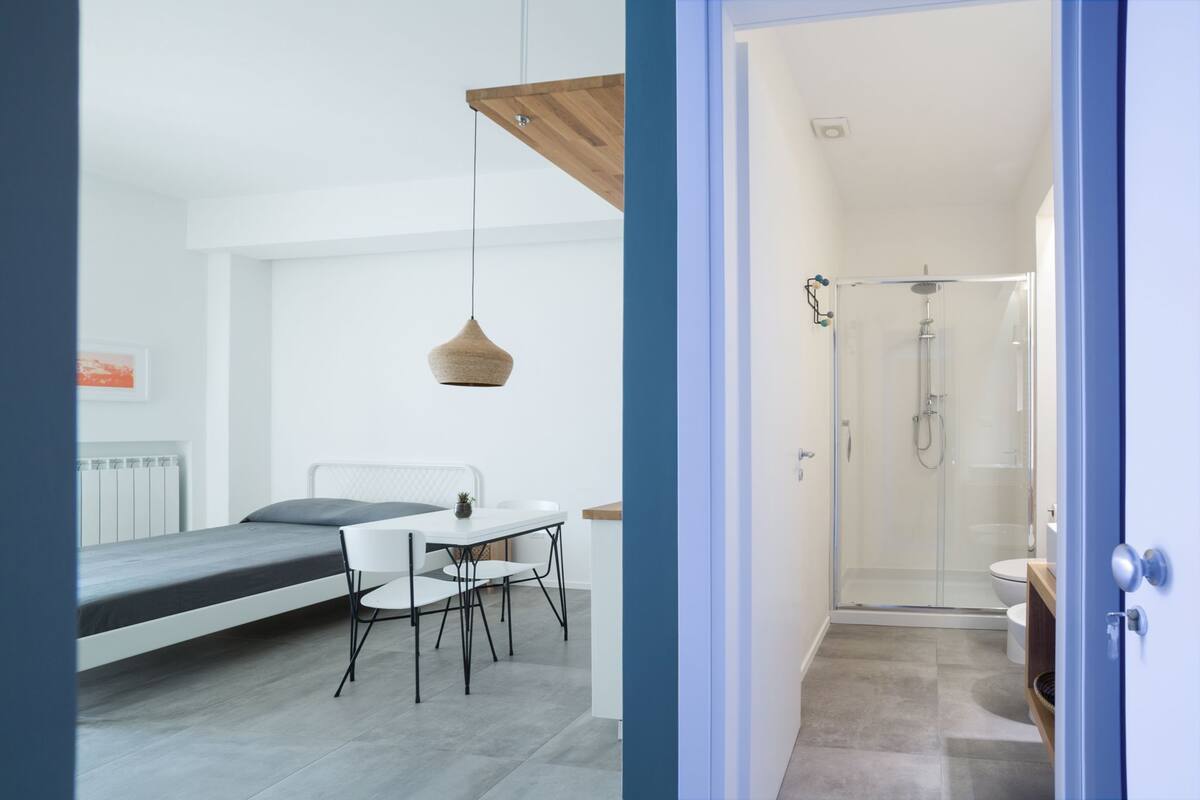 A compact room with a bed, a small table with chairs, a hanging light fixture, and a shower area.