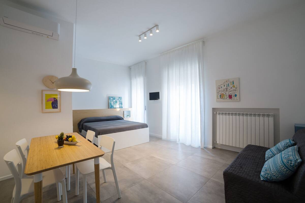 A modern, well-lit room with a bed, a dining table with chairs, a sofa, and a radiator.