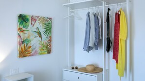 A white closet with hanging clothes, a hat on a small table, and a tropical leaf painting on the wall. - Salsedine Sea View (Salerno)