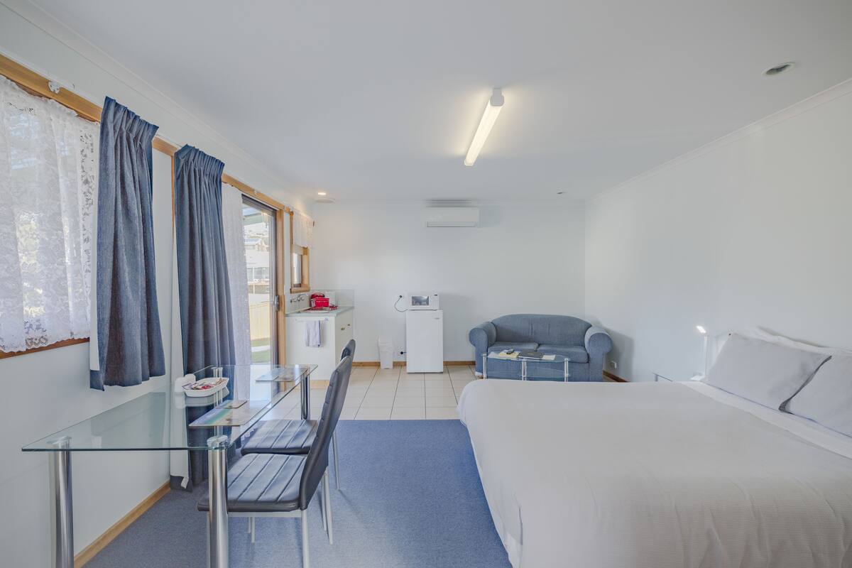 A hotel room with a bed, a desk with a chair, a small kitchenette, and a window with curtains.