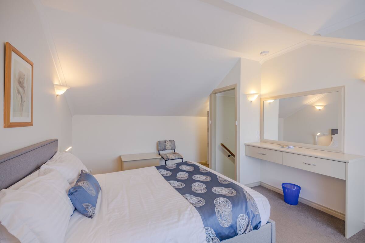 A bedroom with a bed, a chair, a dresser, a mirror, and a wall-mounted light.