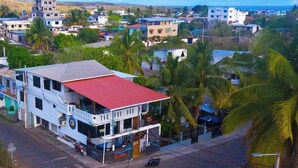 A multi-story building with a red roof, located in a residential area with visible palm trees and other houses. - The Island Home (Puerto Ayora)