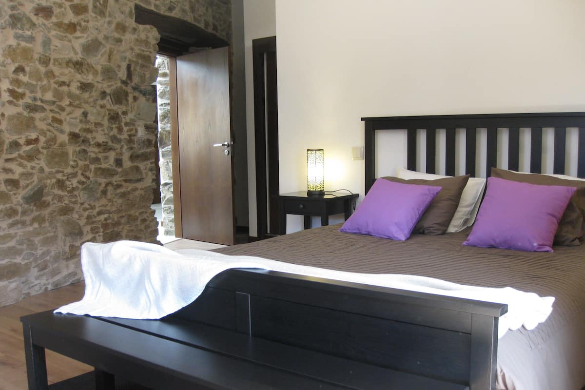 Un dormitorio con pared de piedra, estructura de cama de madera, mesita de noche con lámpara y ventana con vistas a la pared de piedra.