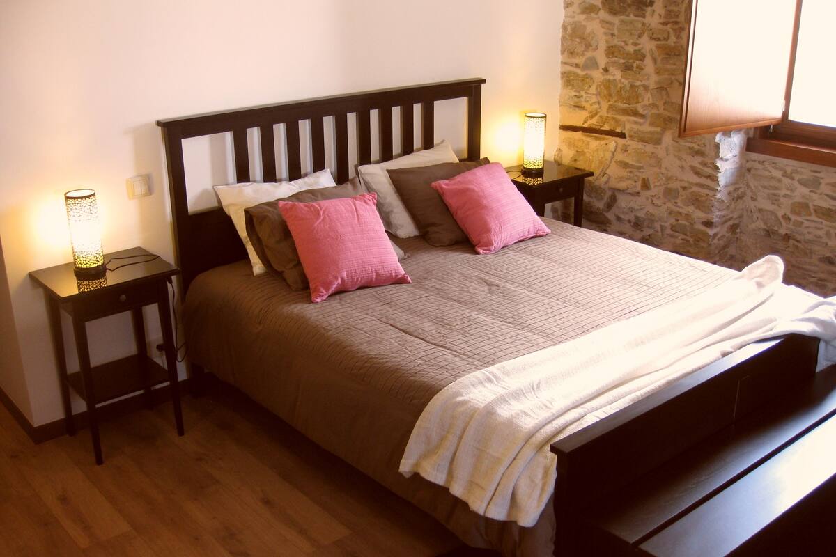 Un dormitorio con cabecera de madera, una cama grande con muchas almohadas, dos mesitas de noche con lámparas, una pared de piedra y una ventana.