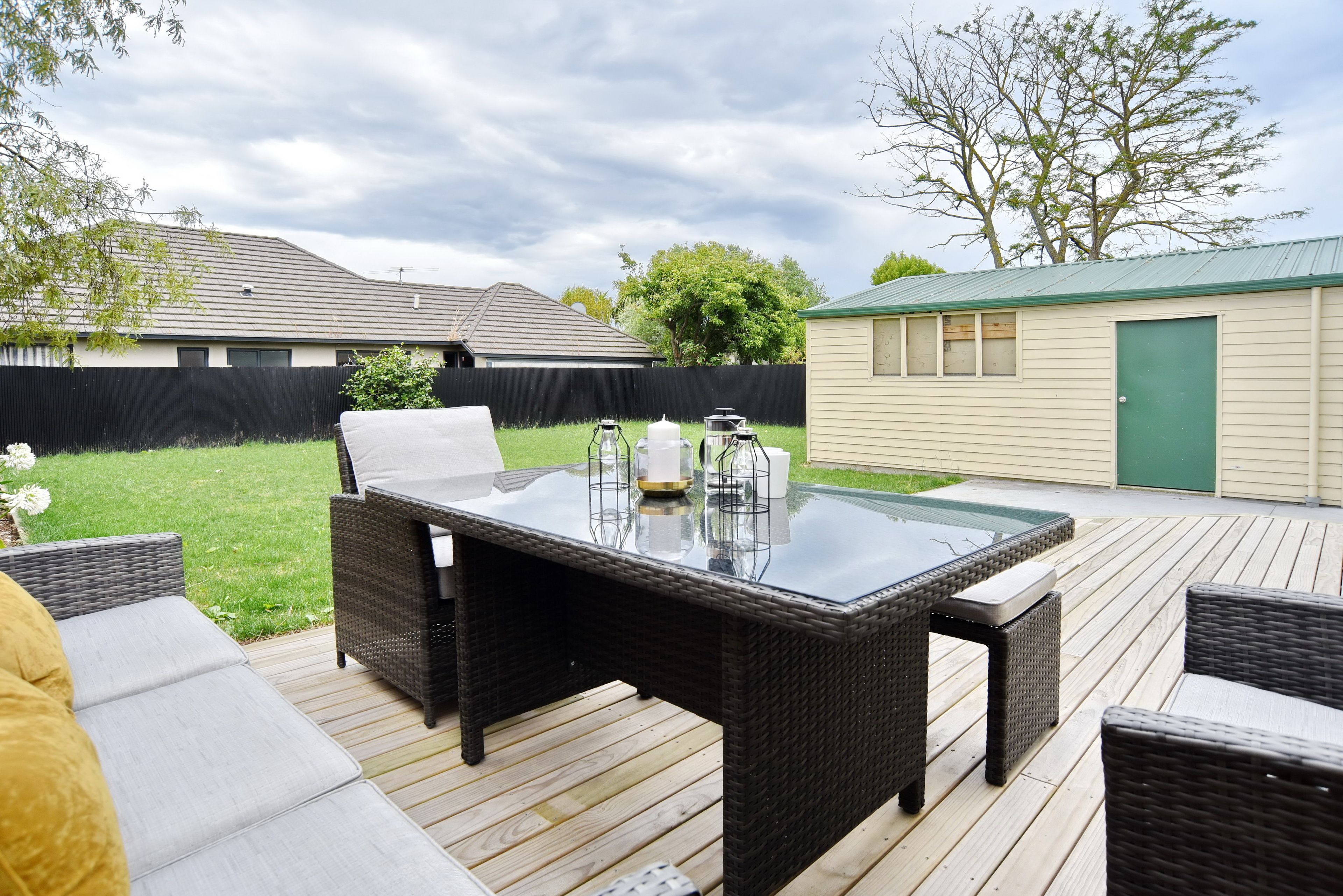 A patio with a glass-top table and wicker furniture, surrounded by a wooden deck and a grassy yard.