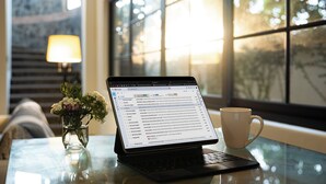 Een laptop op een glazen tafel, met een kopje en een vaas met bloemen.