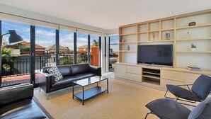 Una sala de estar moderna con un sofá de cuero negro, una mesa de centro de vidrio y un televisor de pantalla plana sobre un soporte de madera. La habitación tiene ventanas grandes que ofrecen vistas a la ciudad.