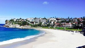 Una playa costera con un litoral rocoso, un mar azul cristalino y una zona residencial con edificios al fondo.