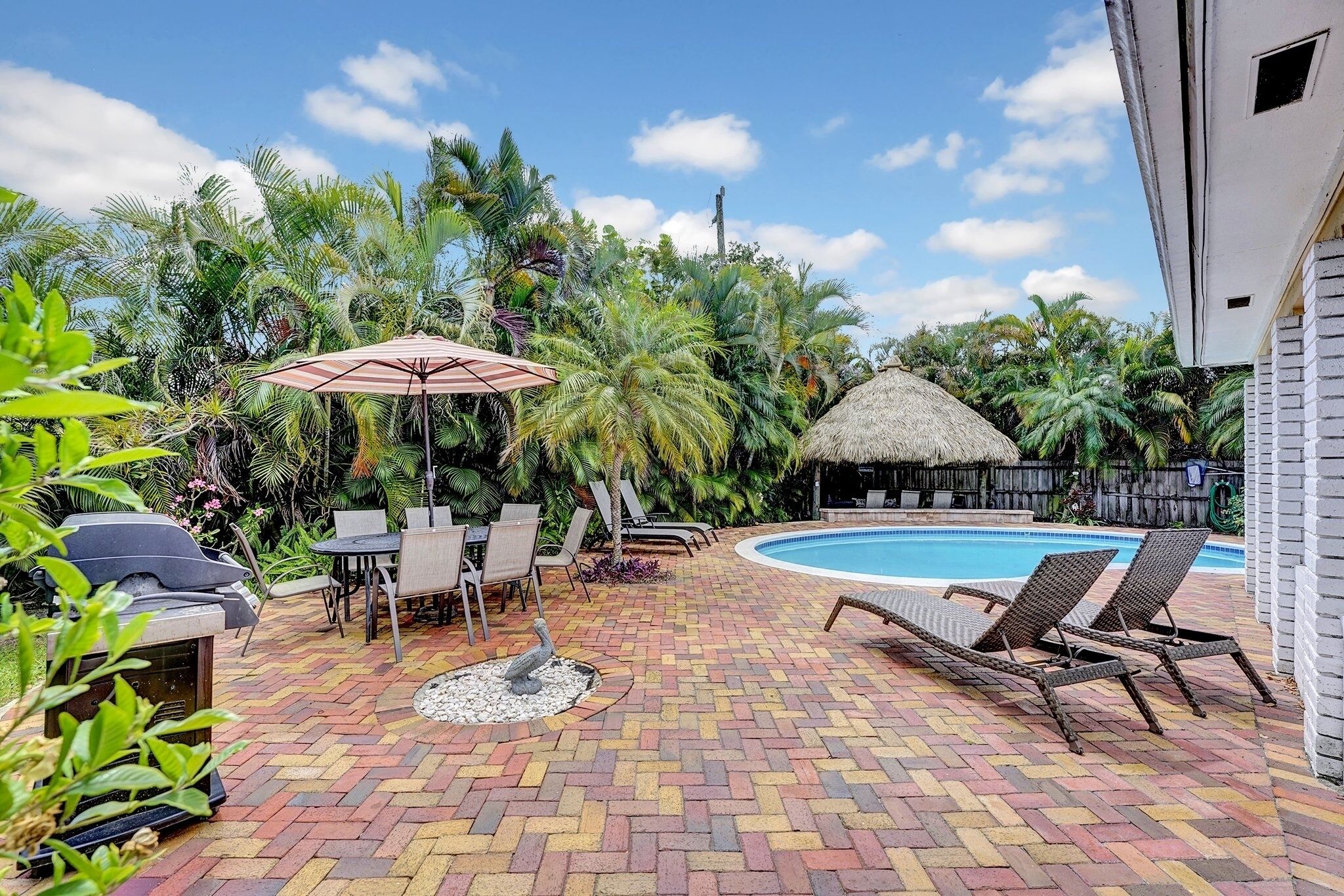 A poolside area with a brick patio, lounge chairs, a grill, and a thatched-roof structure.