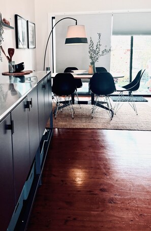 A modern kitchen with a dining area, featuring a wooden floor, black chairs, and a large window with blinds. - Culburra Retreat - Newly Renovated, Pet Friendly, 4 Min Walk to the Beach (Culburra Beach)
