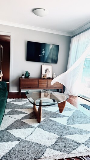 Un salon moderne avec une table basse en verre, un téléviseur fixé au mur et un meuble en bois.
