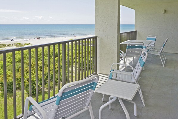 Ein Balkon mit weißen und blauen Stühlen und Tischen mit Blick auf einen Strand, Menschen und den Ozean.