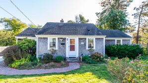 A single-story house with a front porch, a chimney, and a small garden with various plants and flowers.