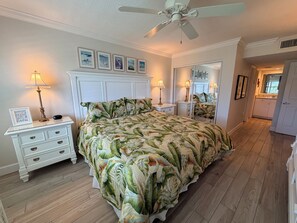 A bedroom with a bed, bedside tables, lamps, a ceiling fan, a mirror, and a hallway leading to a bathroom.