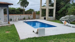 A modern outdoor pool area with a rectangular pool, white lounge chairs, and a small table with chairs. - Apartment in Cabo Frio, completely renovated, new furniture, bright, spacious, airy, quiet. (Cabo Frio)