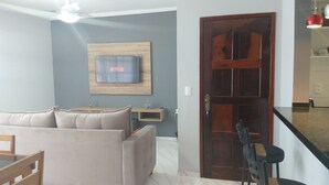 A living room with a sofa, a TV mounted on the wall, a wooden door, and a small table with a water bottle. - Apartment in Cabo Frio, completely renovated, new furniture, bright, spacious, airy, quiet. (Cabo Frio)