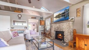 A spacious living room with a stone fireplace, a glass coffee table, and a view of the kitchen in the background. - Sunset Cottage - Waterfront Beach House on Shelter Bay with Private Dock (Southold)