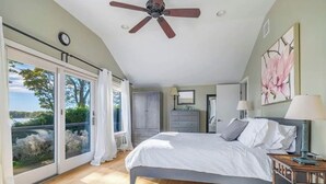 A bedroom with a bed, a ceiling fan, a nightstand, a dresser, and a large window with curtains. - Sunset Cottage - Waterfront Beach House on Shelter Bay with Private Dock (Southold)