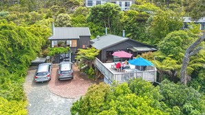 A house with a deck, two cars parked in front, and lush greenery surrounding it. - Coromandel Cottage  - Romantic Rustic - Sea views (Coromandel Township)