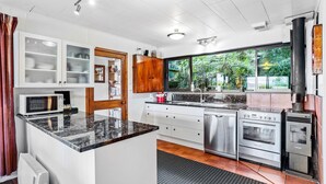 A modern kitchen with white cabinetry, a black granite countertop, stainless steel appliances, and a large window offering a view of greenery outside. - Coromandel Cottage  - Romantic Rustic - Sea views (Coromandel Township)