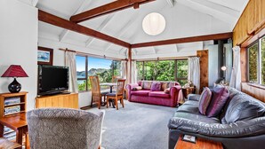A spacious living room with a large TV, wooden furniture, and a view of greenery outside. - Coromandel Cottage  - Romantic Rustic - Sea views (Coromandel Township)