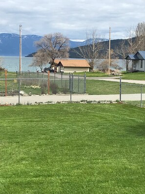 A fenced grassy area with a small building and a dock by a lake, surrounded by mountains.