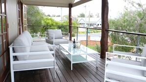 Un espace extérieur aménagé avec des meubles blancs et une table à plateau en verre, protégé par un auvent et offrant une vue sur une rue et des espaces verts.