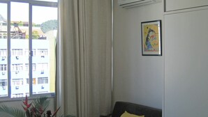 A room with a window, a grey sofa with a yellow pillow, a plant, and an air conditioning unit on the wall.