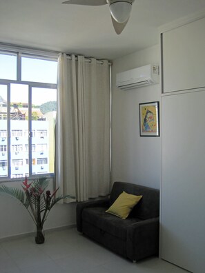 Quarto com janela, sofá cinza com almofada amarela, uma planta e um aparelho de ar condicionado na parede.