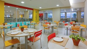 A brightly lit cafeteria with round tables and chairs, potted plants, and large windows offering views of the outside.