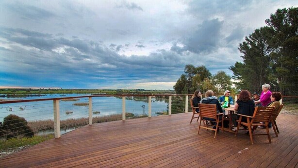 A wooden deck with a group of people seated at a table, overlooking a body of water and a distant landscape. - Limestone Ridge River Getaway (Tailem Bend)