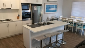 A modern kitchen with white cabinetry, a built-in oven, a refrigerator, and a sink. There is a dining area with white chairs and a table. - Moontana @ Moonta Bay (Moonta Bay)