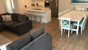 A modern living room with a grey sofa, a small kitchen area with a fridge, oven, and bar stools, and a dining table with chairs. - Moontana @ Moonta Bay (Moonta Bay)
