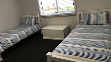 A bedroom with two single beds, a small white bench, and a window with blinds.
