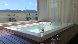 A hot tub on a wooden deck with a glass barrier, a glass of water, and a view of a building and hills in the background. - Spend your holidays at a club. (Nações)
