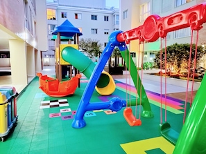 A children's play area with slides and swings, surrounded by a building with balconies and windows.