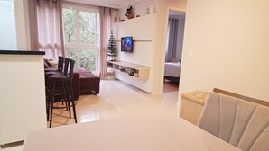 A modern living room with a white sofa, a dining table with chairs, a TV mounted on the wall, and a small Christmas tree. - Spend your holidays at a club. (Nações)