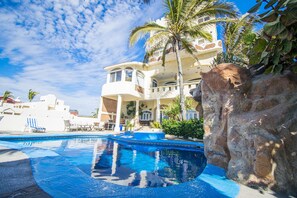 A pool area with a clear blue pool, lounge chairs, and a large building with balconies and palm trees.