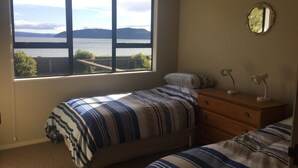 A bedroom with a large window offering a view of a body of water and hills. There is a bed with striped bedding and a dresser with two lamps. - Stunning beach front bach (Kinloch)