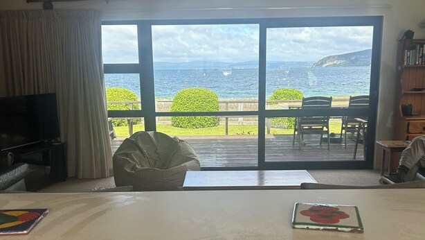 A room with a large sliding glass door leading to a balcony with a view of the sea. There is a television on the left, a table with a placemat, and a bookshelf on the right. - Stunning beach front bach (Kinloch)