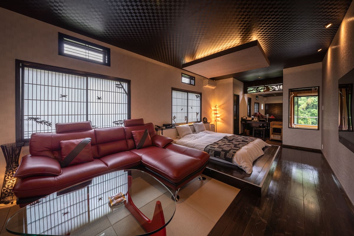 Een moderne hotelkamer met een groot bed, een rode leren bank, een glazen salontafel en grote ramen met uitzicht op groen.