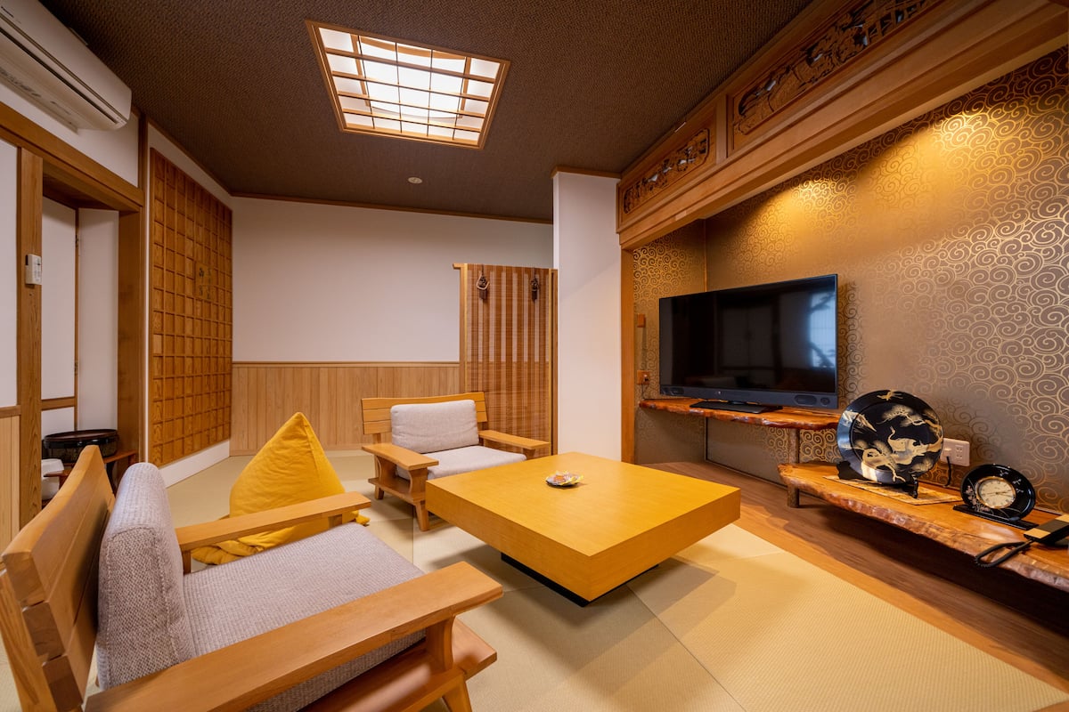 Een traditionele Japanse woonkamer met tatami vloerbedekking, een lage houten salontafel, een bank met kussens, een aan de muur bevestigde televisie en een decoratief zwart-wit bord op een houten plank.