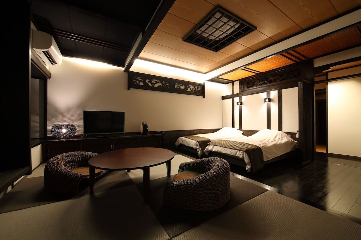 Een traditionele Japanse kamer met tatami vloeren, een lage tafel, twee zachte zitplaatsen, een bed met een wit matras en bruin beddengoed, en een televisie op een standaard.