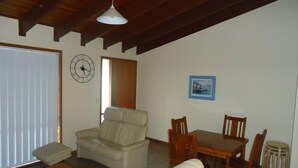 A living room with a sofa, a dining table with chairs, a clock on the wall, and a door leading to another room.