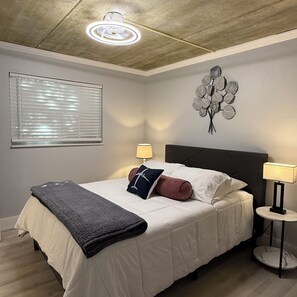Ein Schlafzimmer mit einem Bett, zwei Nachttischlampen, einer Wandleuchte, einem Fenster mit Jalousien und einem Wanddekor.
