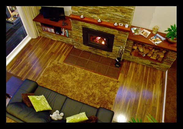 Ein Wohnzimmer mit Kamin aus Stein, einer Couch mit Kissen, einem Fernseher auf einem Holzregal und einem kleinen Tisch mit Zeitschriften und einer Pflanze.