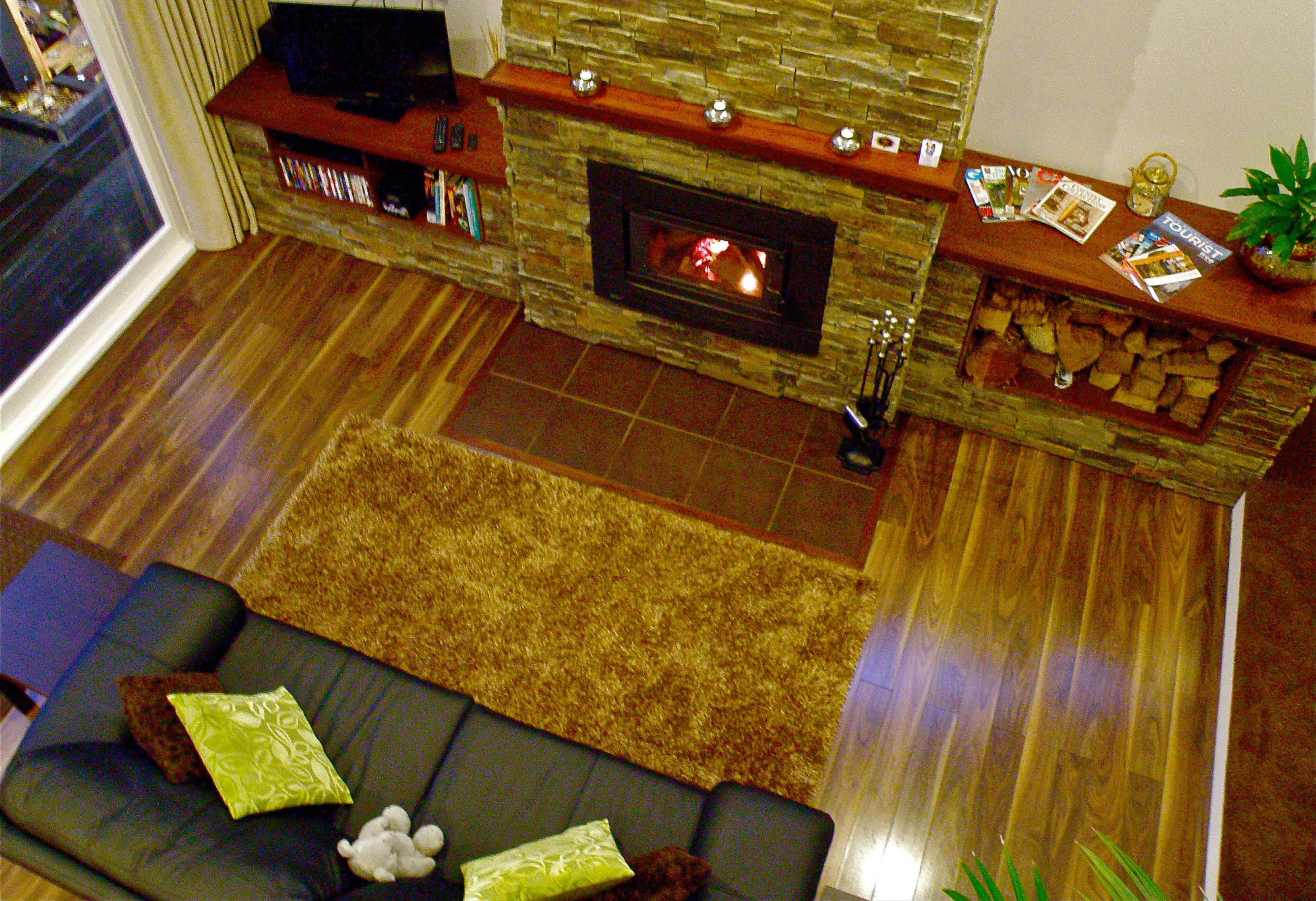A living room with a stone fireplace, a sofa with cushions, a television on a wooden shelf, and a small table with magazines and a plant.