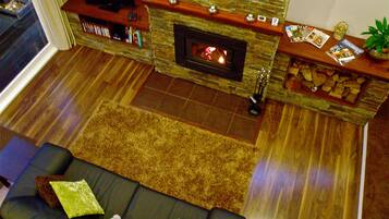 A living room with a stone fireplace, a sofa with cushions, a television on a wooden shelf, and a small table with magazines and a plant.