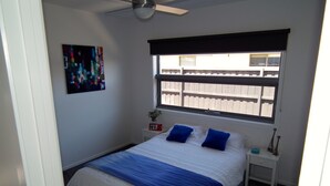 Una camera da letto con un letto, un comodino, una lampada da tavolo, una ventola a soffitto, una finestra vista un recinto in legno e un quadro appeso al muro.