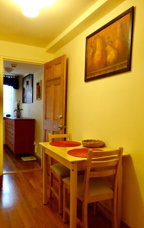 A dining area with a table and chairs, featuring a painting of fruits on the wall and a cabinet in the background.